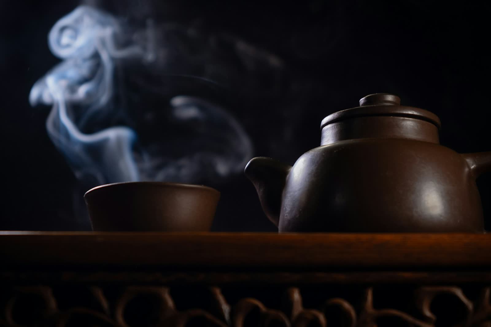 A steaming kettle/teapot and cup sitting on a dark tabletop, wisps of steam catching the low light.